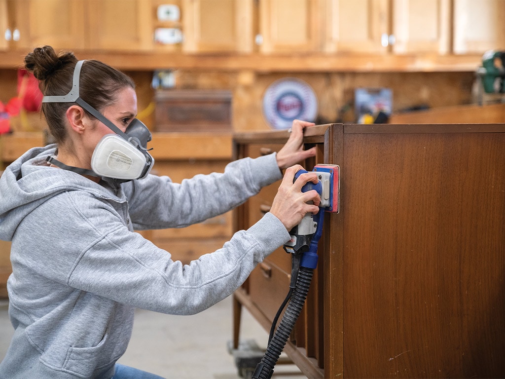 Jana Strangler wearing a mask using a sander on a dresser