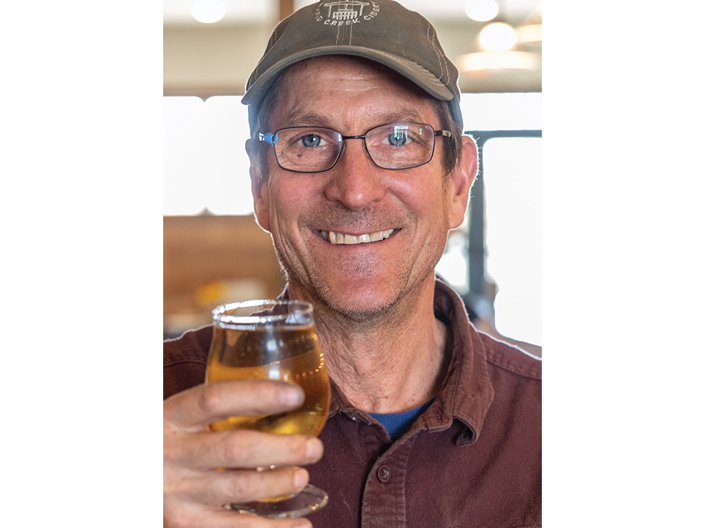 closeup of smiling man wearing cap and glasses, holding a apple cider in a glass