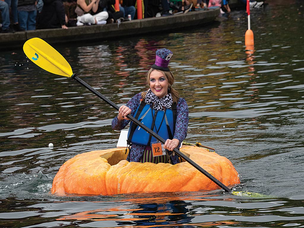 a woman in a costume and top hat rowing a giant carved out orange pumpkin