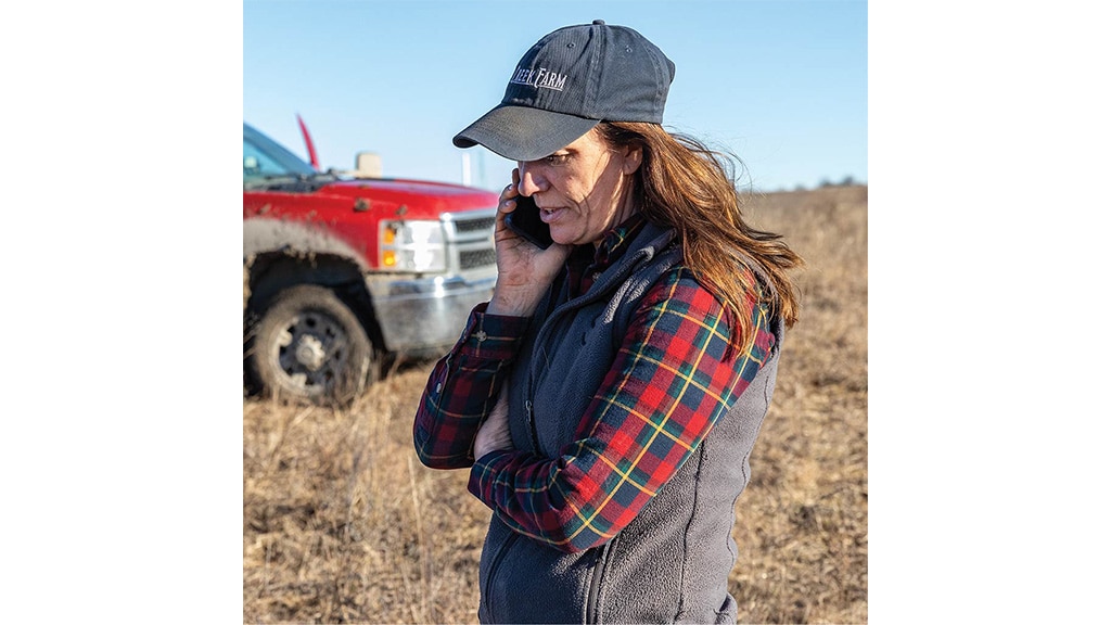 Person holding mobile phone to face with baseball cap, long brown hair, red plaid shirt, grey vest with red truck in a field in an out of focus background