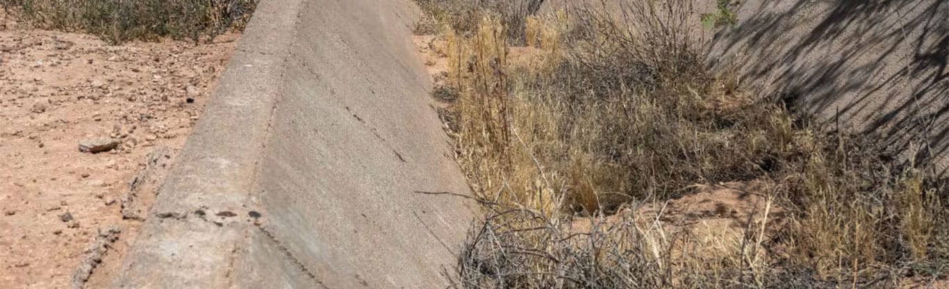 A concrete drainage ditch in the desert full of brown shrubs