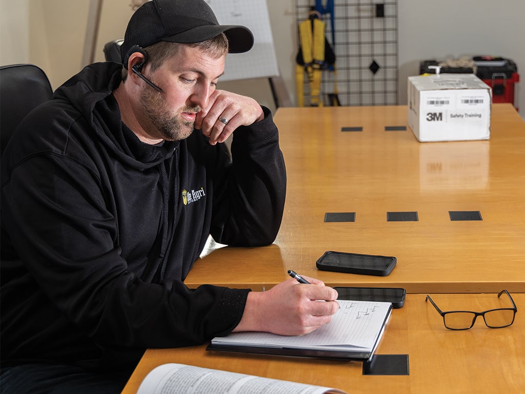 Person at a table writing in a notebook with a smartphone, glasses, and a 3M box in the background.