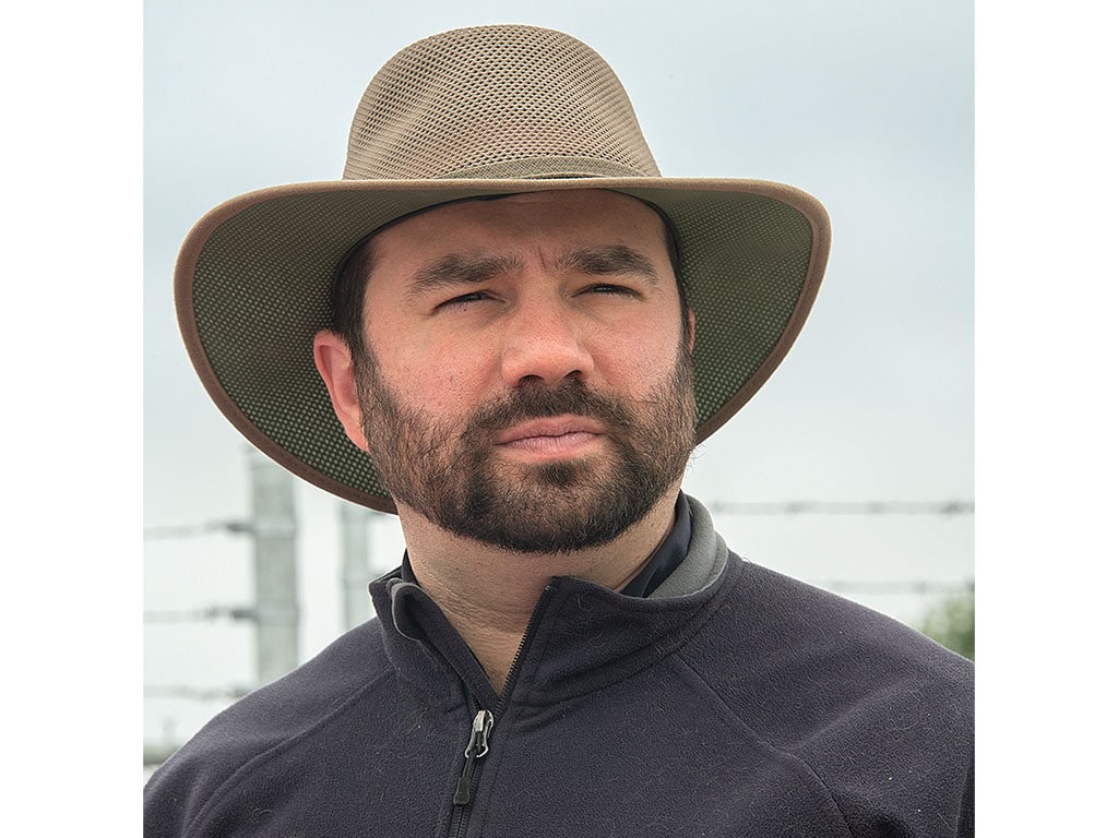 A person wearing a tan wide-brimmed hat and a dark zip-up jacket stands outdoors, with power lines and greenery in the background.