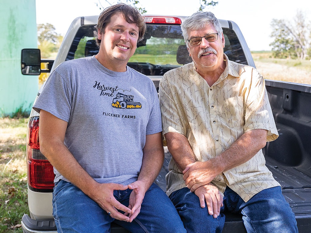 Two men sitting on the tailgate of a pickup truck and smiling.