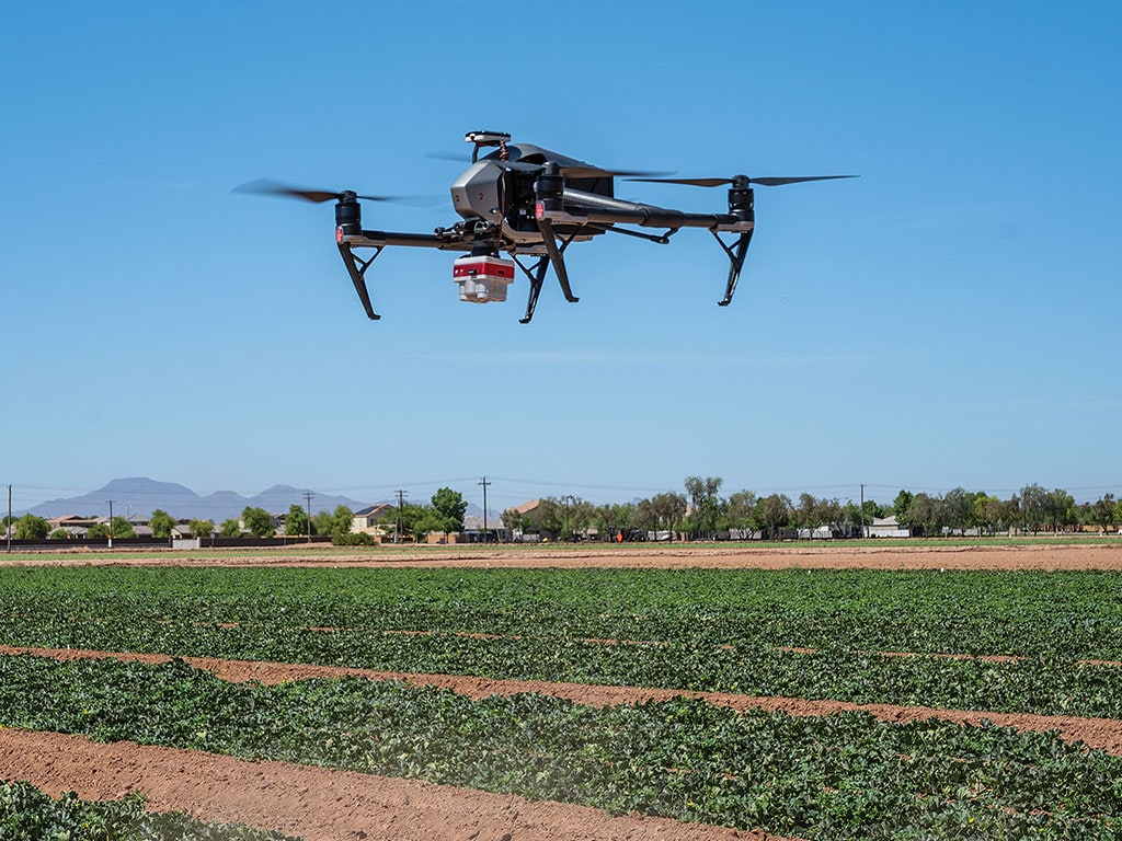 Agricultural drone flying over a green crop field with mountains in the background.