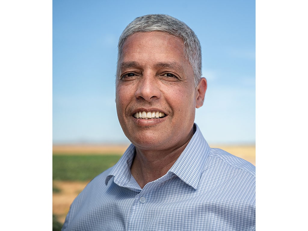 Man in checkered shirt standing outdoors with a blue sky and field background.