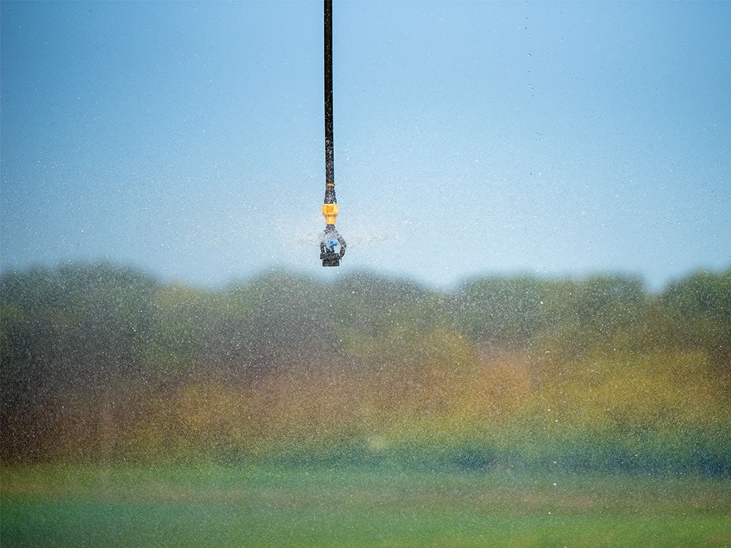 A sprinkler head spraying water droplets in the air with a blurred green landscape in the background.