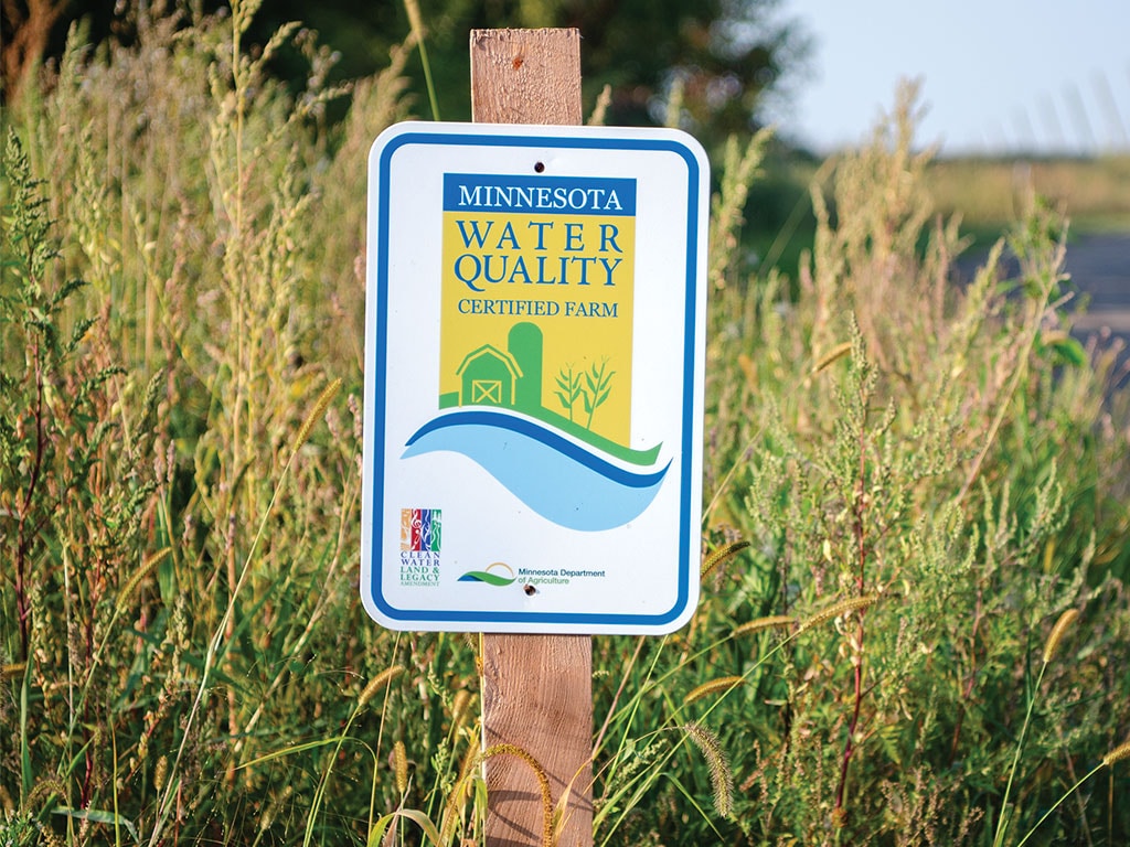 A wooden sign reading "Minnesota Water Quality Certified Farm" stands among tall grasses, promoting sustainable farming practices.