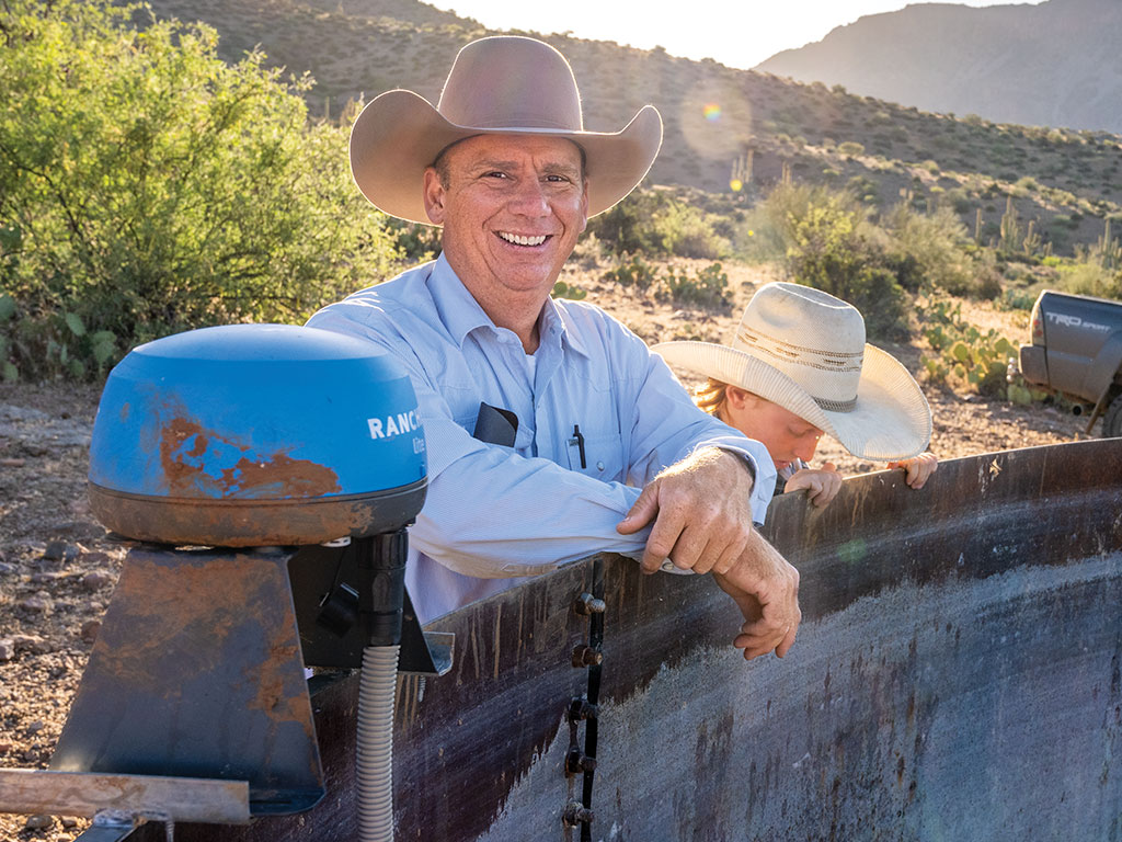 Two individuals in cowboy hats leaning on a metal trough in a sunny desert landscape.