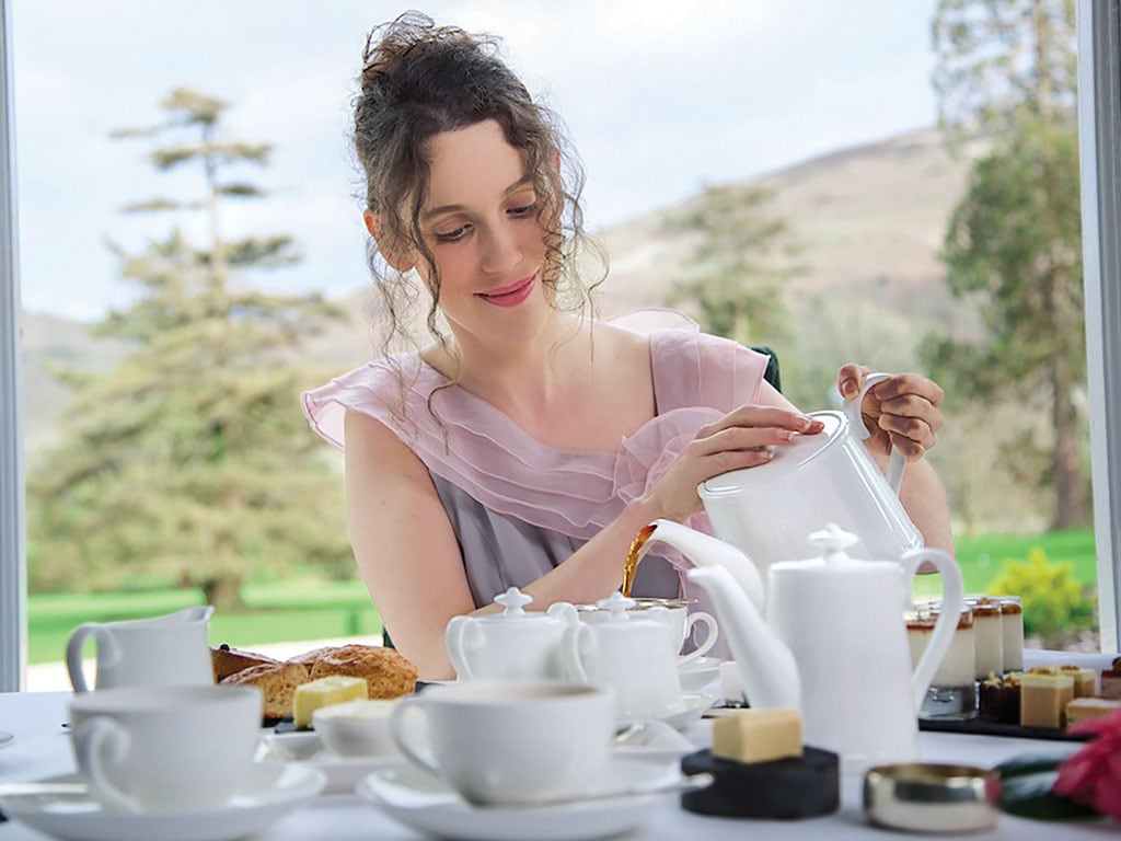 Person pouring tea at a table with cups and cakes, with a scenic backdrop.