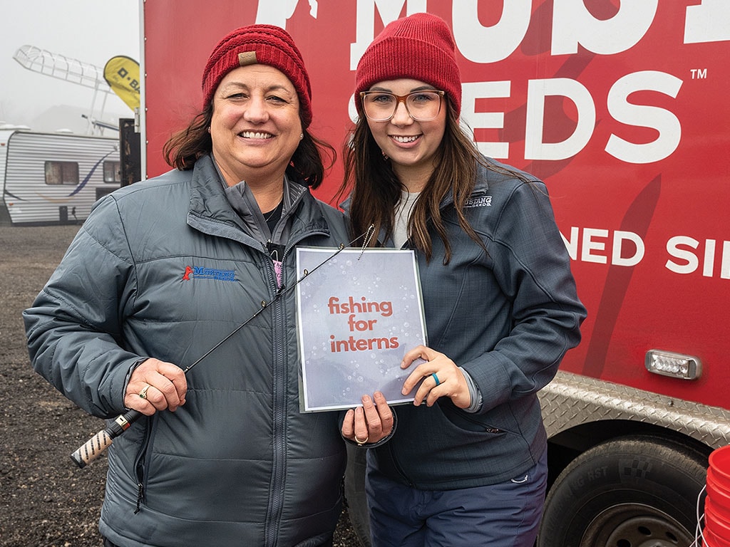 Two individuals holding a sign stating "fishing for interns" next to a red truck.