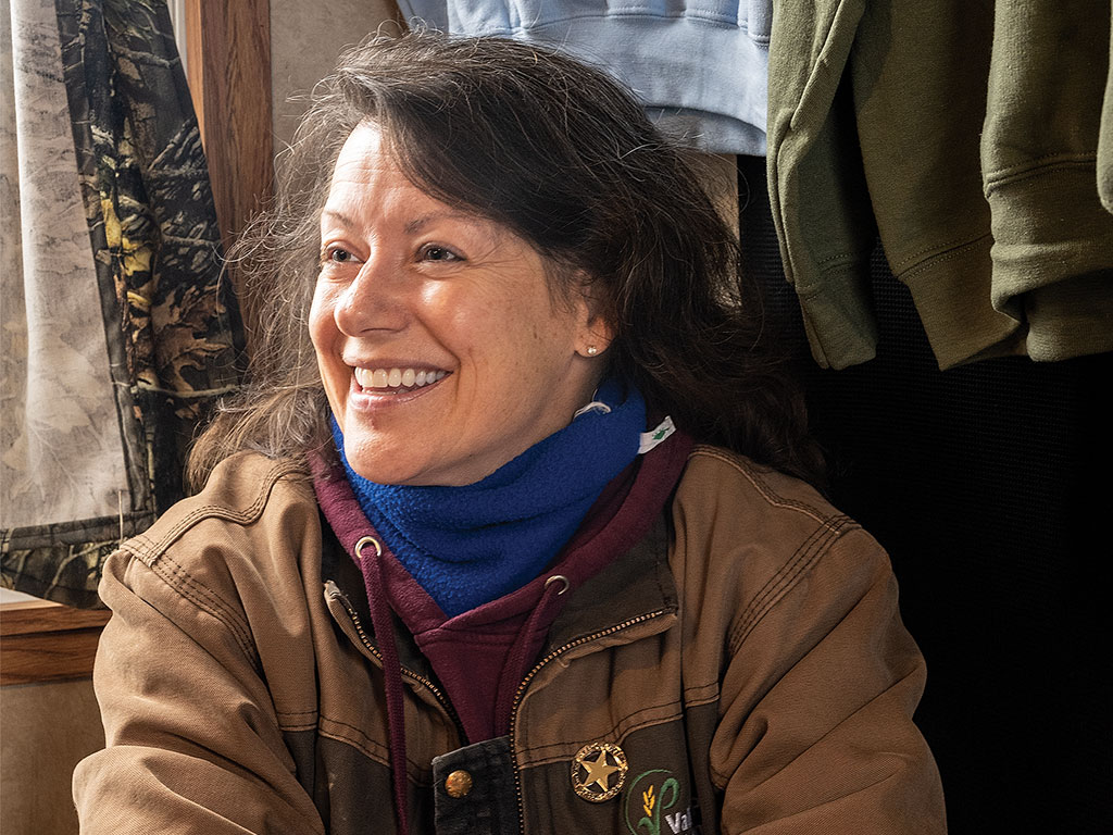 Person in a brown jacket sitting indoors with camouflage drapery and clothing in the background.