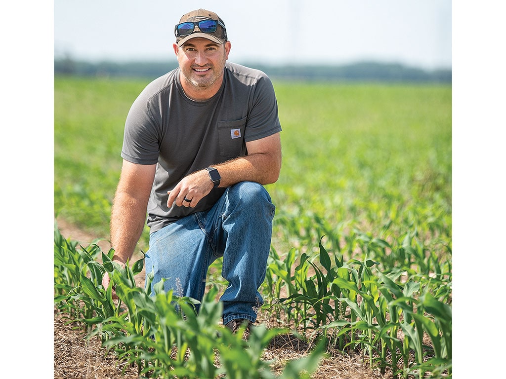 Man in a gray t-shirt and jeans kneeling in a cornfield.