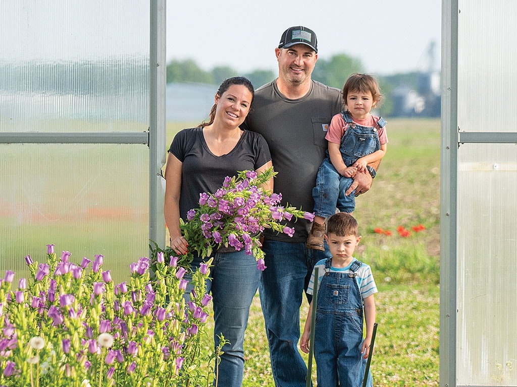 A family of four stands outside a greenhouse, with one adult holding a bunch of purple flowers.