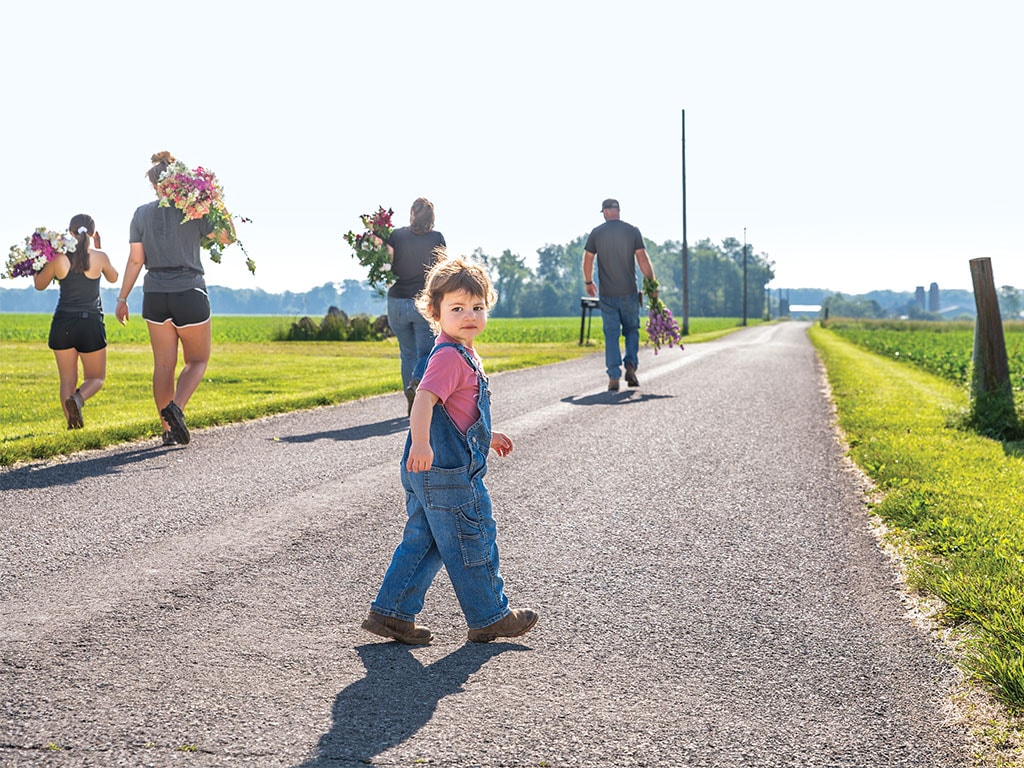 Child in overalls walking on a sunny road with adults carrying flowers ahead.