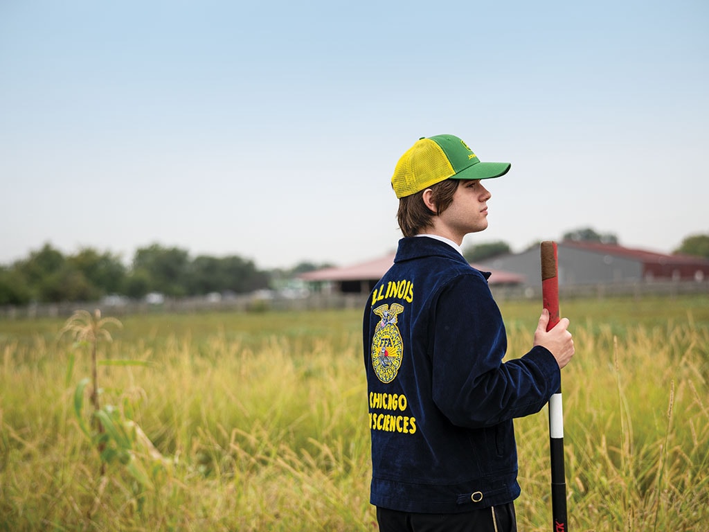 A young person in a blue jacket and yellow hat stands in a field, holding a shovel.