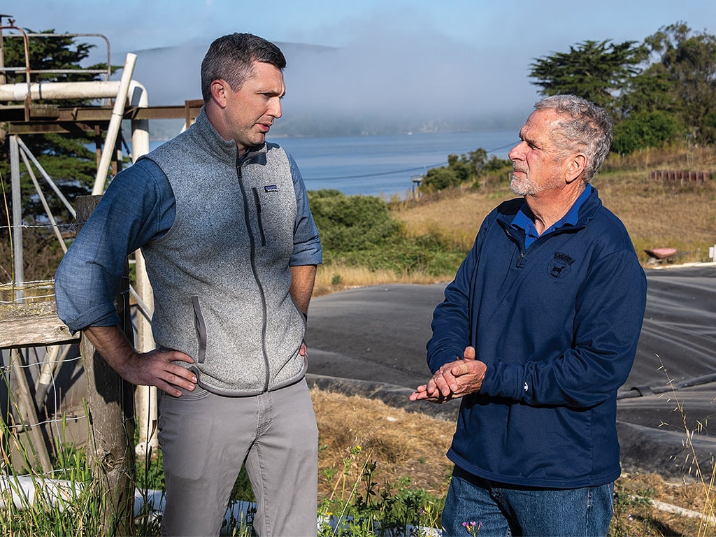 Two men standing outdoors with a scenic backdrop of hills and water.