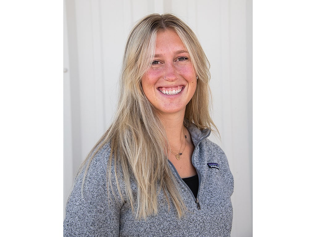 Person with long blonde hair wearing a grey zippered jacket against a white door.