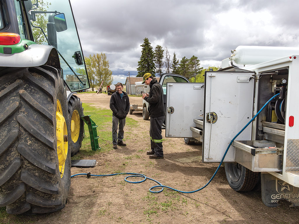 Two individuals near a tractor and a fuel tank, with a hose connecting them, in a rural setting.