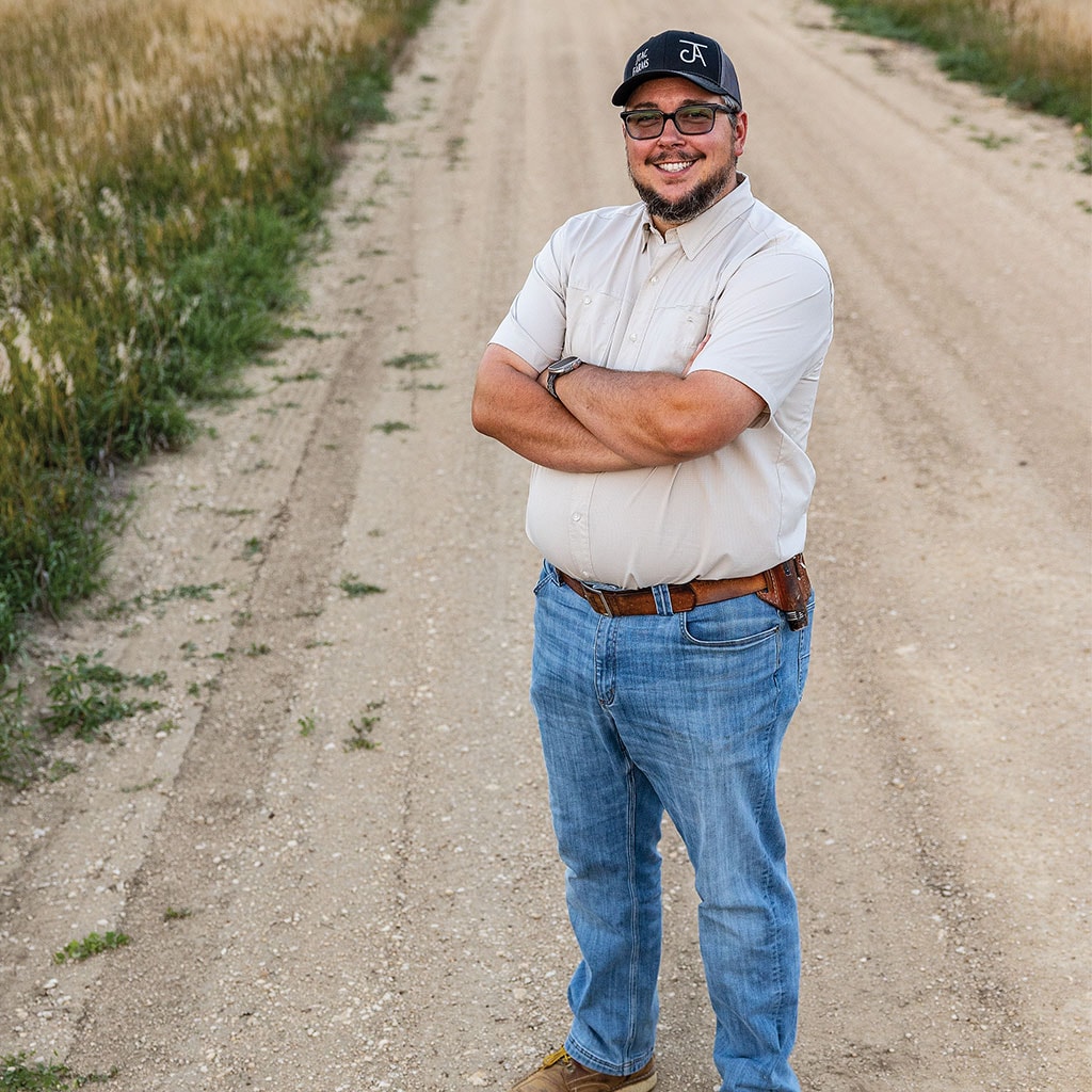 Man standing with arms crossed on a dirt road next to a field.