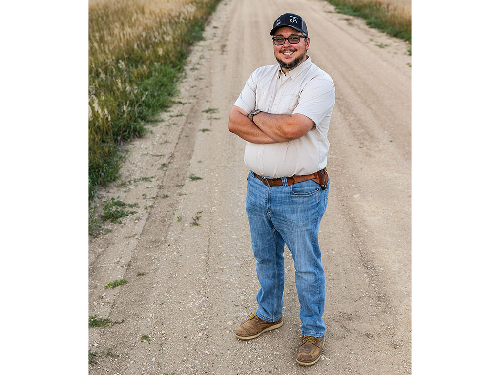 Man standing with arms crossed on a dirt road next to a field.