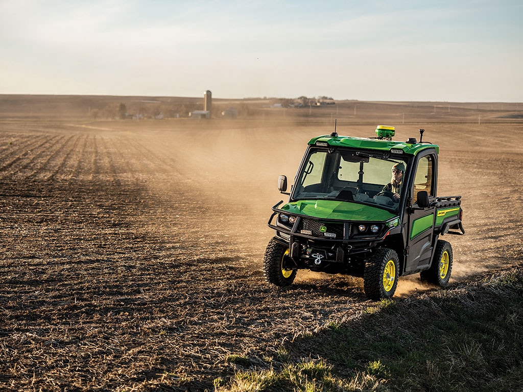 Utility vehicle drives over a dry, dusty field with a farm and silo in the distant background.