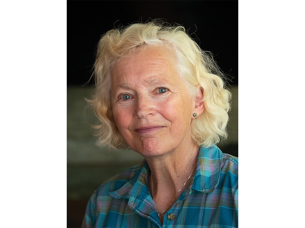 A smiling blonde-haired person with a teal, purple and yellow plaid collared shirt on