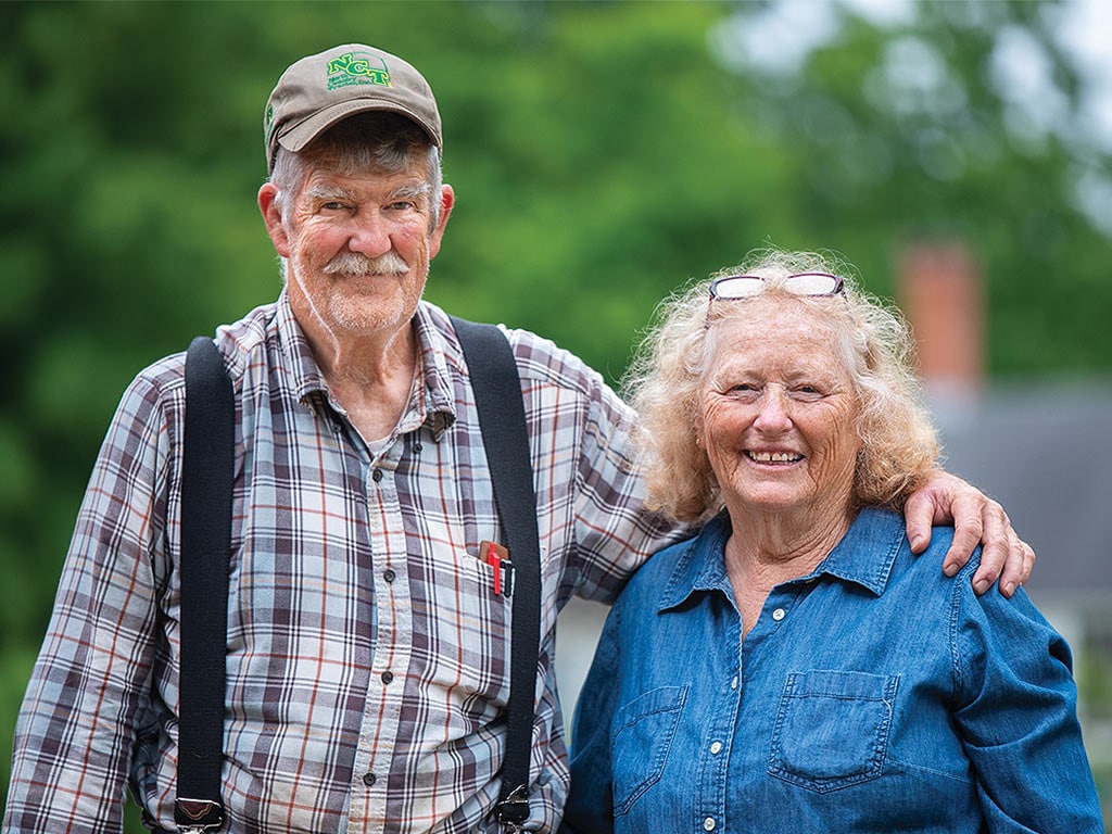 A smiling elderly couple stands together outdoors, wearing casual clothing with a blurred background of trees and a house.