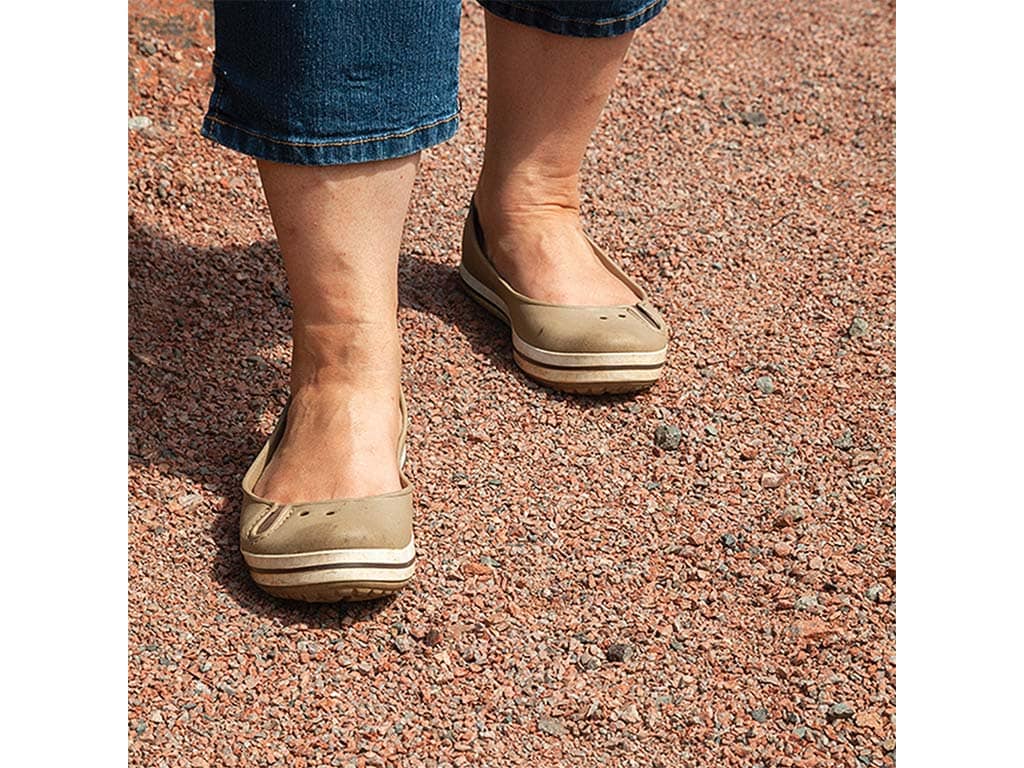 Close-up of a person's feet in beige flat shoes, standing on rocky, reddish gravel soil, wearing rolled-up denim jeans.