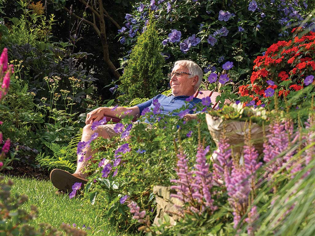 A person relaxes on a garden bench surrounded by vibrant flowers, including purple, pink, and red blooms, creating a serene atmosphere.