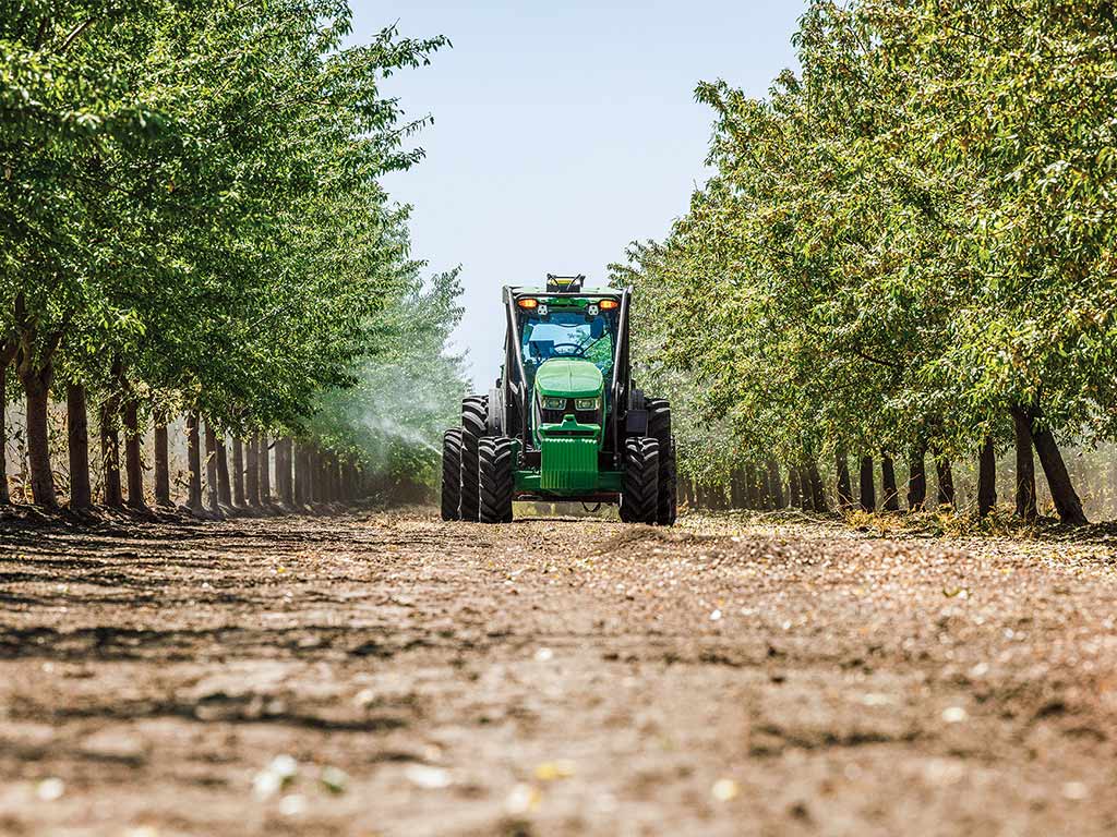 A driverless John Deere tractor driving on a dirt path between two rows of trees