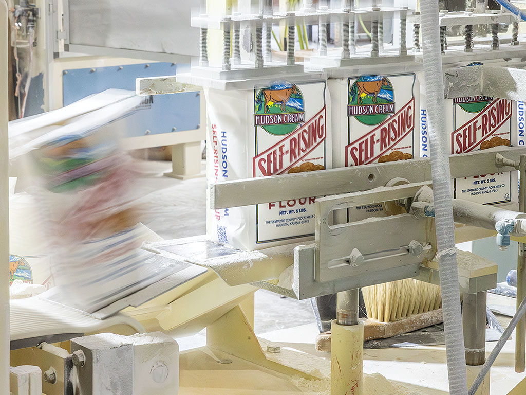 A packaging machine processes Hudson Cream self-rising flour bags on a conveyor, with flour dust visible in the air.