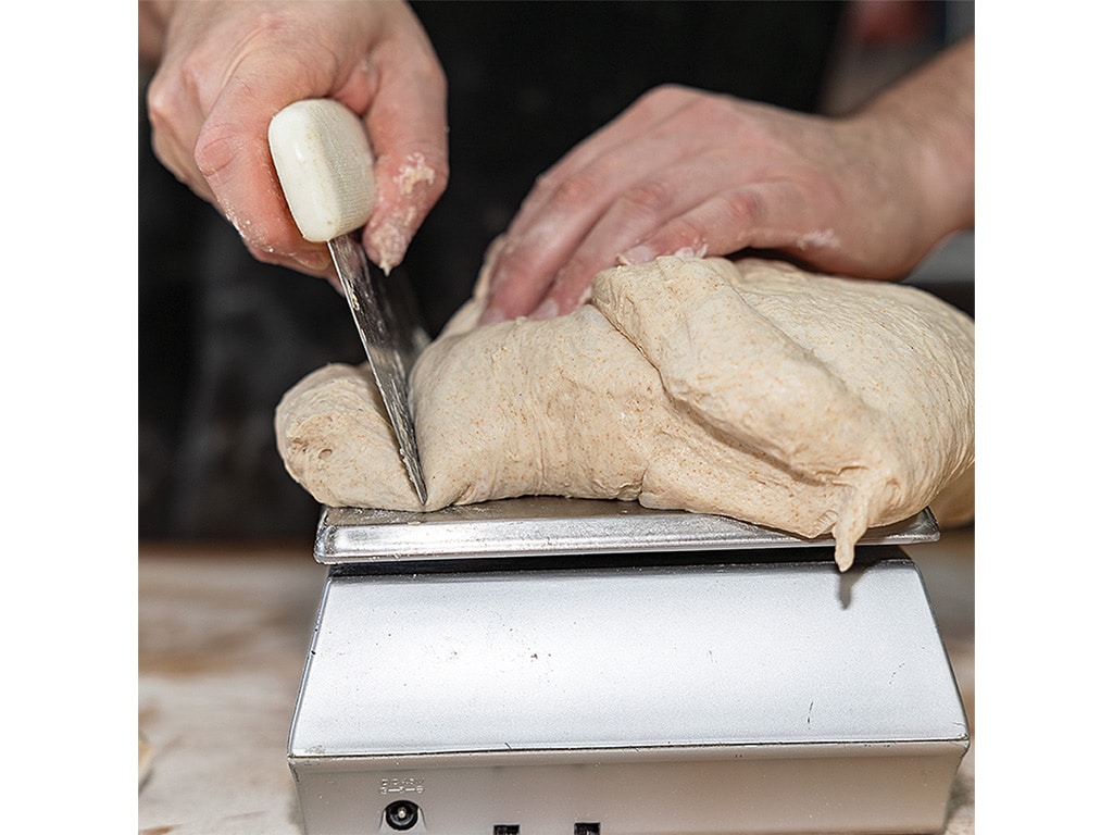 A baker uses a knife to cut a lump of dough resting on a silver scale, preparing to weigh it for baking.