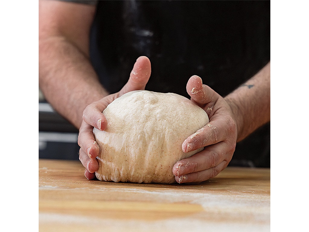 A baker's hands knead a round ball of dough on a wooden surface, dusted with flour, ready for baking.