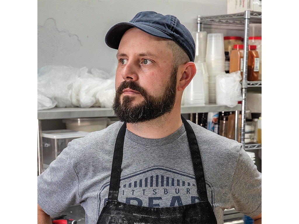 A man wearing a cap and a gray t-shirt stands confidently in a kitchen filled with various cooking supplies and ingredients.