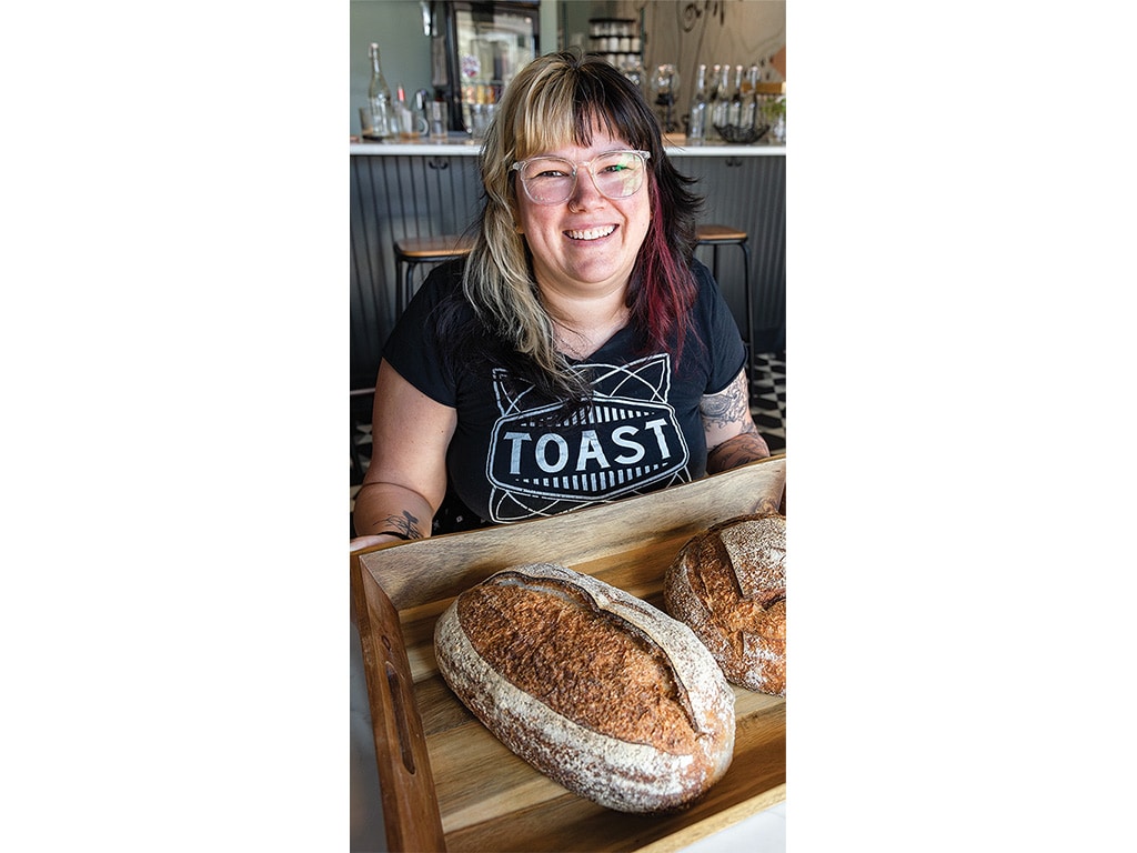 A person in a black "TOAST" shirt holds a wooden tray with two artisanal loaves of bread against a café backdrop.