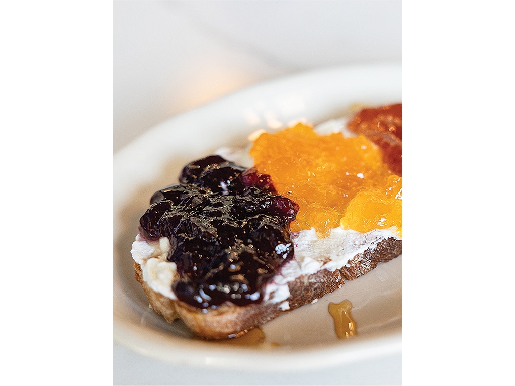 A slice of brown bread topped with cream cheese and three vibrant jams: blackberry, orange, and a golden jelly, drizzled with honey.