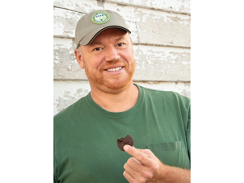 A closeup of a farmer wearing a baseball hat holding a cookie and smiling.