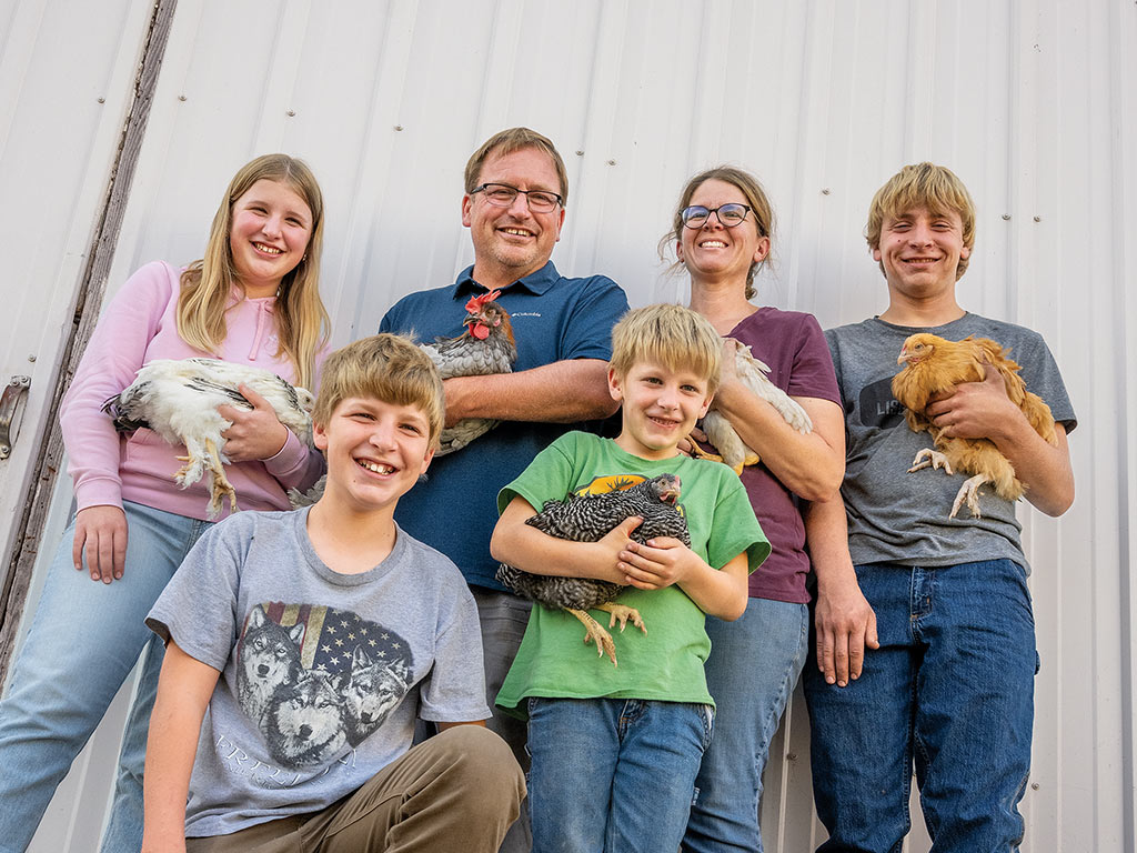 Chicken farming family each holding a chicken standing in front of a corrugated white metal wall