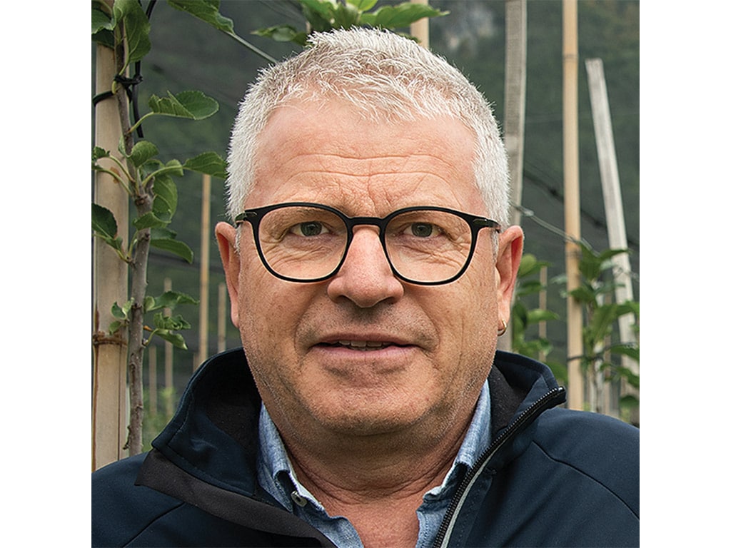 A man with gray hair wearing a black jacket stands in a orchard filled.