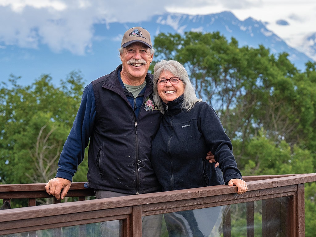 A couple smiles together on a wooden deck with lush greenery and majestic mountains in the background under a cloudy sky.