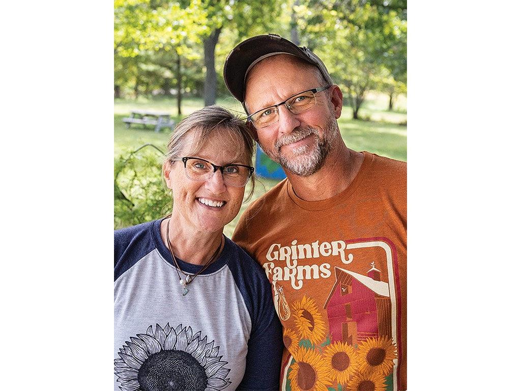 A woman in a sunflower-printed shirt and a man in a vintage farm shirt pose together outdoors, surrounded by greenery.