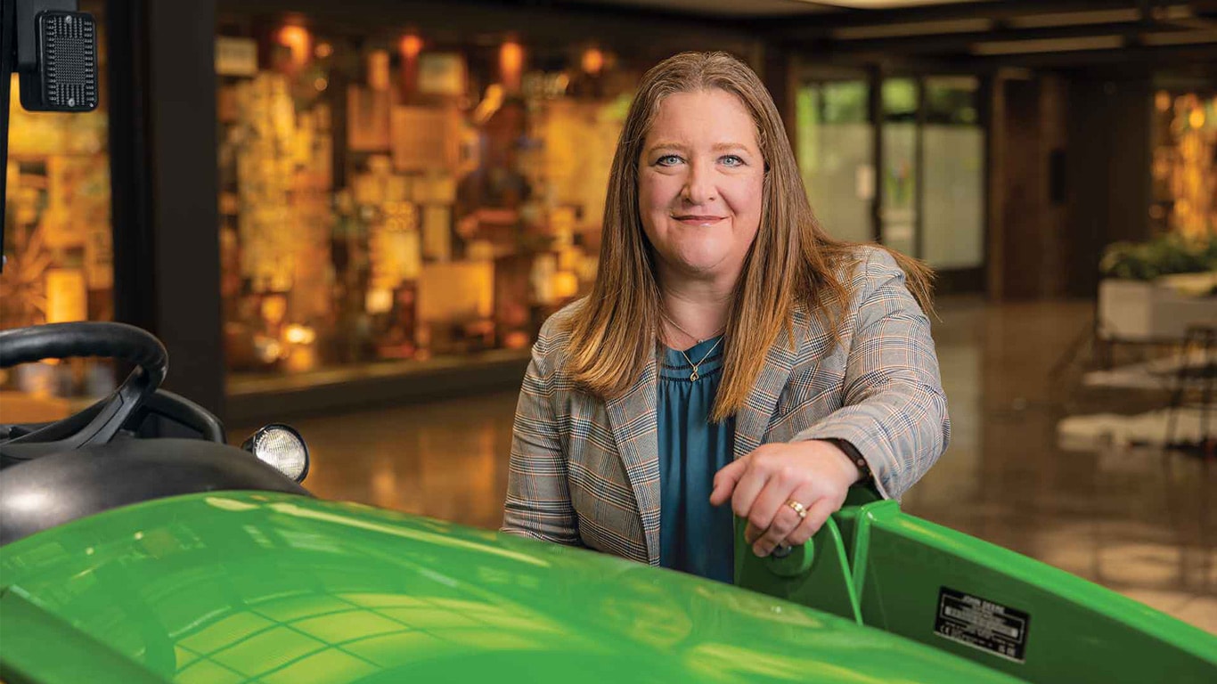 Person standing behind a green tractor in an indoor showroom with displays in the background.