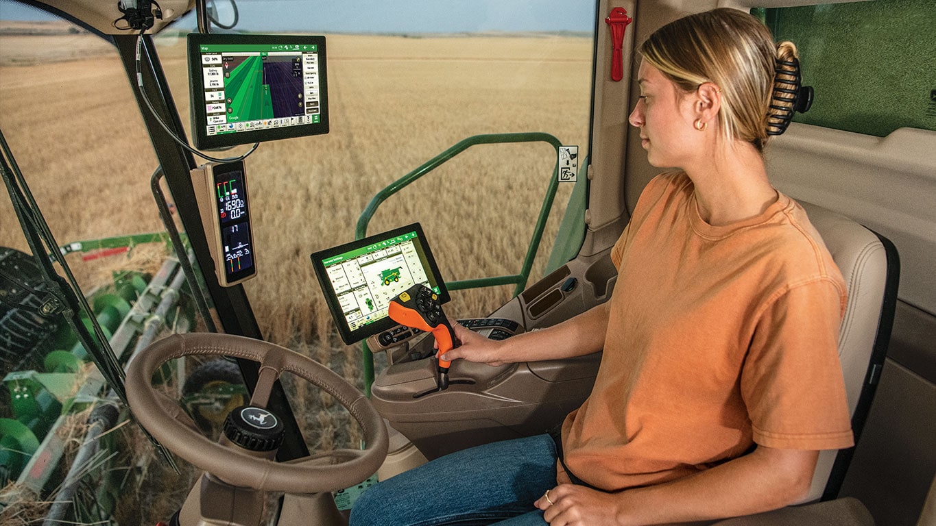 A woman sits in a tractor cab, operating G5 Advanced displays and controls, surrounded by golden harvesting fields.